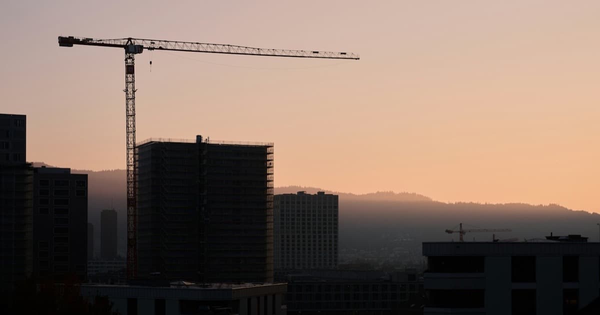 A tower crane silhouetted against a dusk sky over a skyline of buildings under construction.