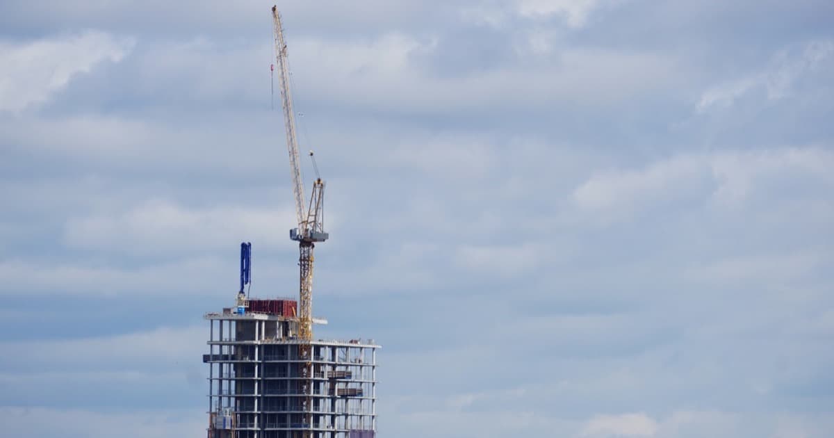 A high-rise building under construction with a crane at its top against a cloudy sky.