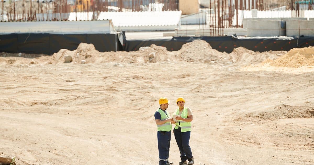 Two site workers in hard hats and hi-vis vests conferring on an earthworks site.